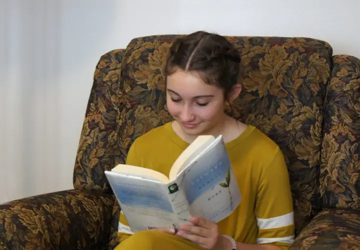 Dziewczyna w warkoczach czyta książkę "Between Shades of Gray", idealną dla 14 latki. Siedzi w wygodnym, wzorzystym fotelu.