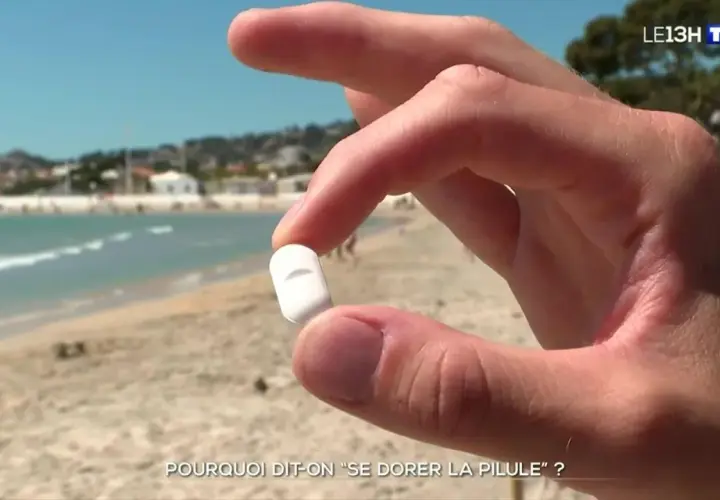 Main d'un homme tenant une pilule blanche sur une plage ensoleillée, interrogeant sur l'expression "se dorer la pilule".