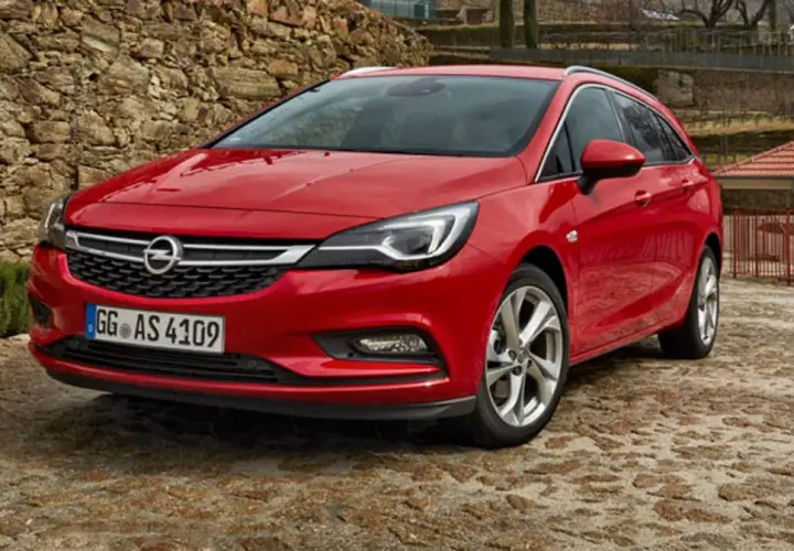 Czerwony Opel Astra Sports Tourer zaparkowany na kamienistej nawierzchni, z kamiennym murem w tle.