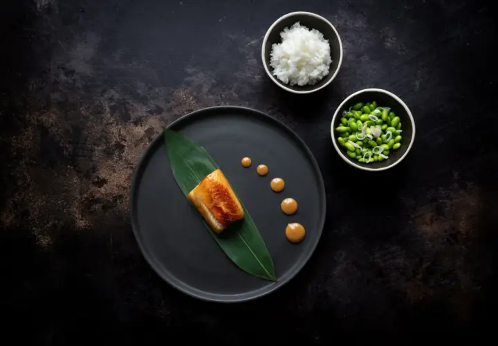 A perfectly cooked nobu miso black cod rests on a dark plate with a green leaf. Drizzled sauce and sides of rice and edamame complete the elegant dish.