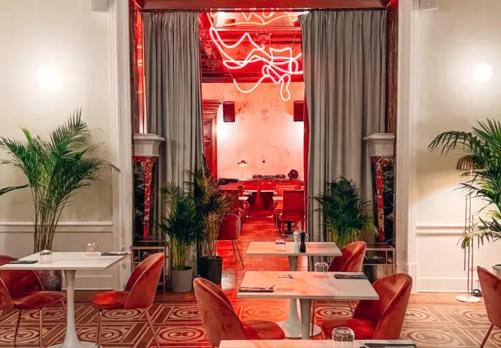 Eleganckie wnętrze z czerwonymi aksamitnymi krzesłami i marmurowymi stolikami. Neonowy napis w głębi sugeruje, że to najdroższa restauracja w Warszawie.