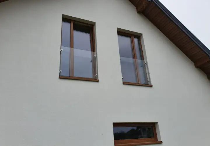 Nowoczesny portfenetr z drewnianymi ramami i szklaną balustradą na białej elewacji.