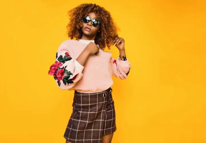 Mujer con cabello rizado y gafas de sol posa con un suéter rosa con rosas bordadas y una falda a cuadros. Su estilo es un armario minimalista moderno.