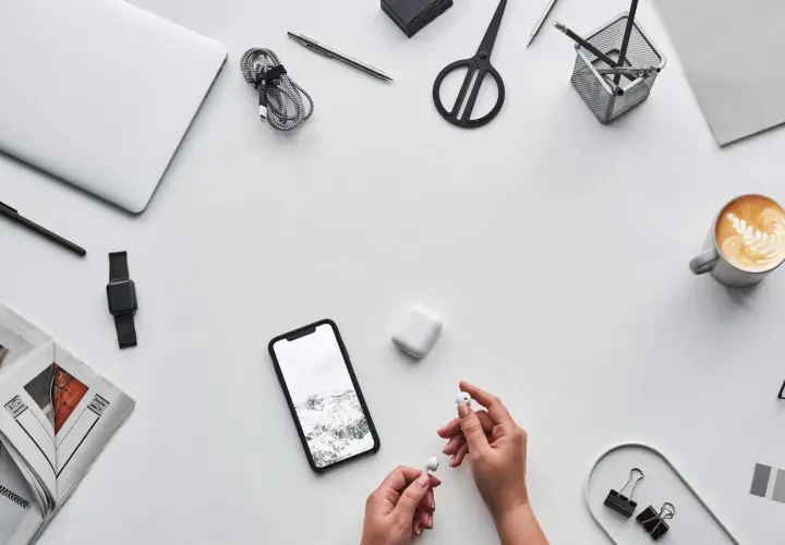 Ręce trzymające słuchawki AirPods obok telefonu z krajobrazem, sugerując, czy airpods działają z androidem. Na biurku laptop, kawa, nożyczki.