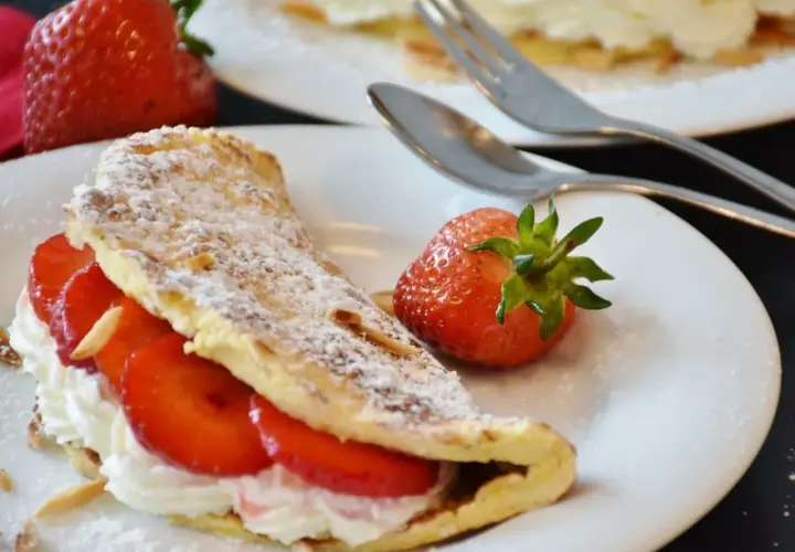 Puszysty omlet z bitą śmietaną i truskawkami, posypany cukrem pudrem i płatkami migdałów. Obok leży świeża truskawka.