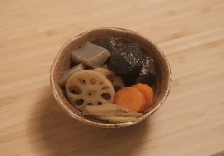 A bowl of simmered vegetables, including shiitake mushrooms, lotus root, carrots, and konjac, likely part of a mushroom dashi broth.