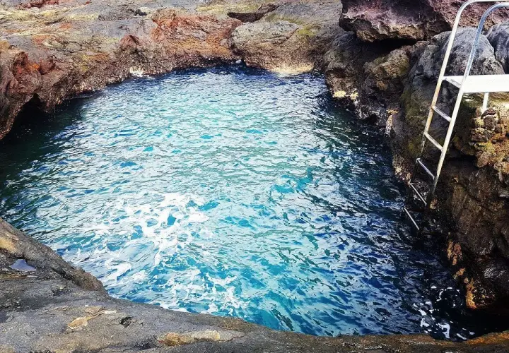 Un charco de los abrigos natural, con aguas cristalinas y una escalera metálica para acceder. Rodeado de rocas volcánicas.