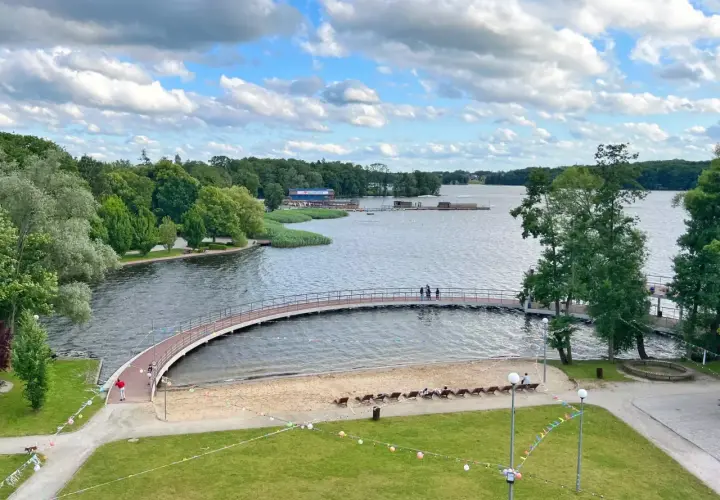 Malownicze jezioro w Szczecinku z łukowatym mostkiem, piaszczystą plażą i zielonymi terenami rekreacyjnymi.