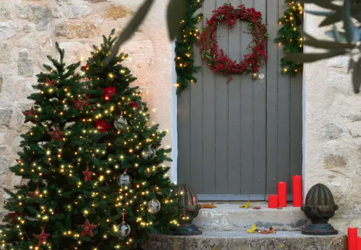 Árbol de Navidad iluminado y una corona de acebo adornan la entrada de una casa de piedra.