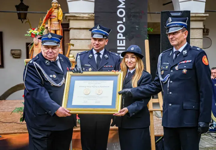 Członkowie OSP otrzymują odznaczenie. W tle drewniana figura i baner.