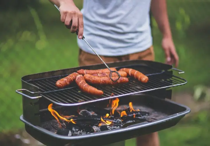 Un homme retourne des saucisses sur un barbecue bois ou charbon. Les flammes dansent sous la grille, promettant un repas savoureux.