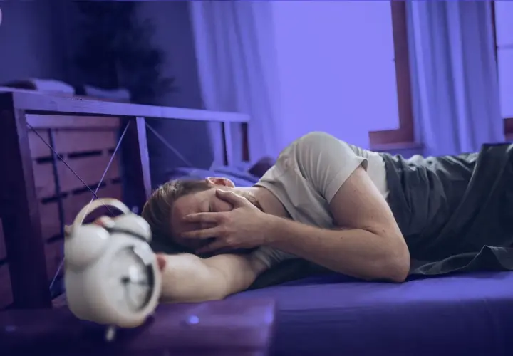 Hombre en la cama, con la cara entre las manos, junto a un despertador. Parece que me levanto cansado.