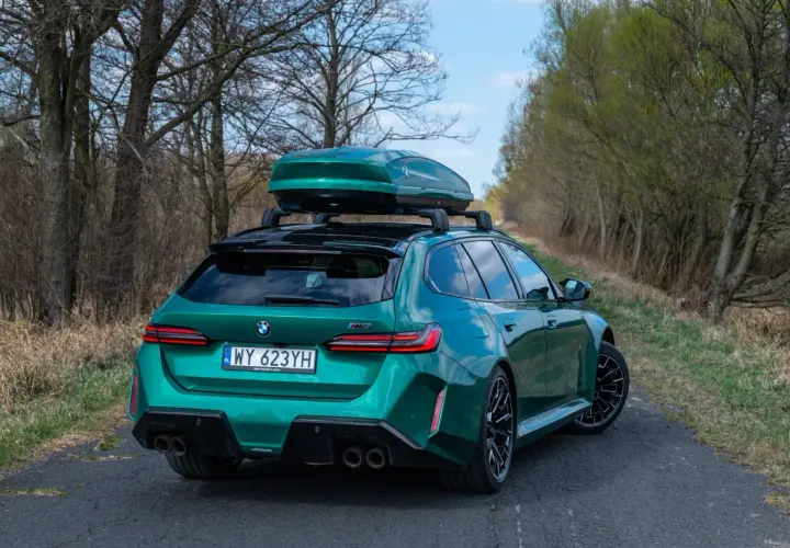 Zielone BMW M5 Touring z boxem dachowym na tle drzew i nieba.