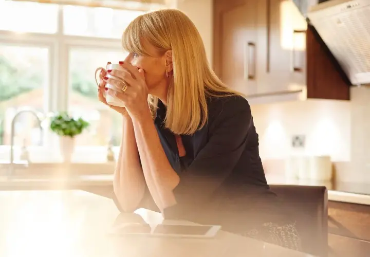 Donna bionda con tazza di caffè riflette sulla sua crisi di mezza età, con luce solare che filtra in cucina.
