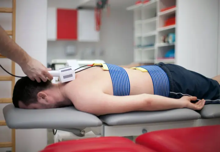 Pacjent leży na kozetce, poddawany zabiegowi z użyciem prądów Traberta. Elektrody przymocowane są do jego pleców.