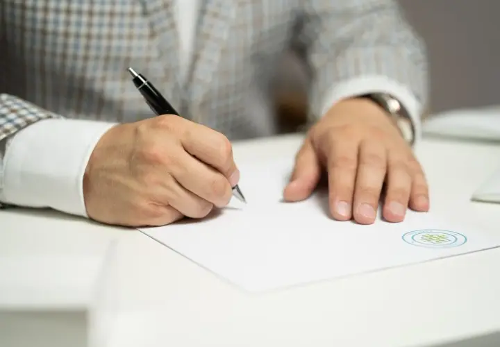 Mężczyzna w eleganckim garniturze pisze długopisem na białej kartce papieru.