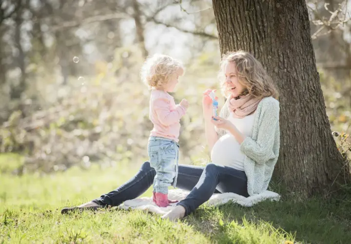 Ciężarna kobieta i małe dziecko puszczają bańki mydlane pod drzewem. Mama uśmiecha się, wiedząc, że robi wszystko, żeby cię chronić.