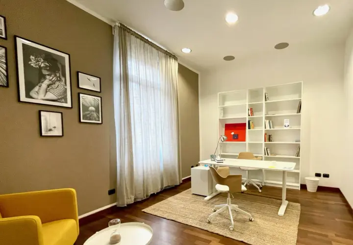 Arredamento studio psicologo: scrivania bianca, libreria, sedie, poltrona gialla, quadri e tenda.