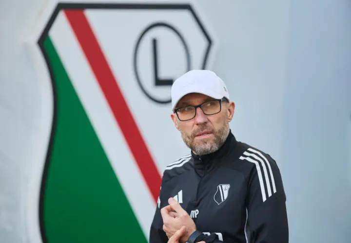 Nowy trener Legii w białej czapce i okularach, na tle klubowego herbu.
