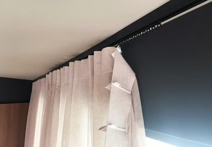 Jasne, lniane zasłony z białymi klipsami, zawieszone na czarnej szynie sufitowej. Eleganckie fleksy do firan zapewniają idealne ułożenie materiału.