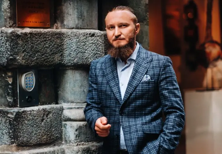 Elegancki mężczyzna w garniturze w kratę, z brodą, oparty o kamienną ścianę. Idealny przykład, jaki krawat do garnituru w kratę wybrać.