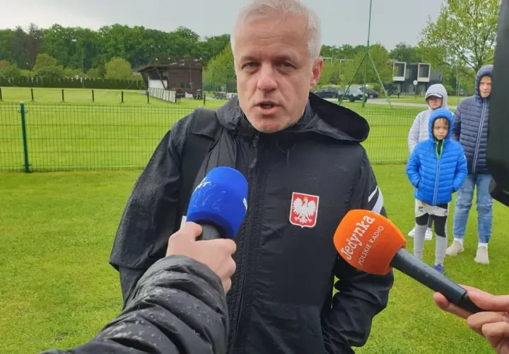 Jacek Jaroszewski, w czarnej kurtce z orłem, udziela wywiadu na boisku. W tle młodzi piłkarze.