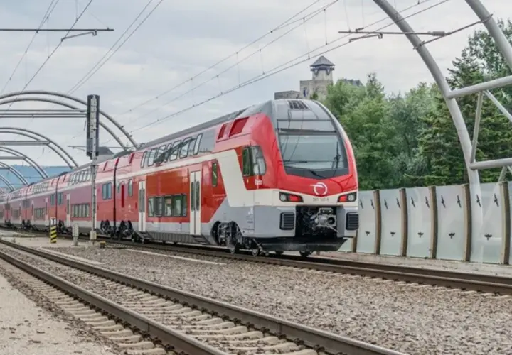 Moderner Doppelstockzug der **Slovak Railways** fährt unter einer Bogenkonstruktion auf Gleisen. Im Hintergrund eine Burg.
