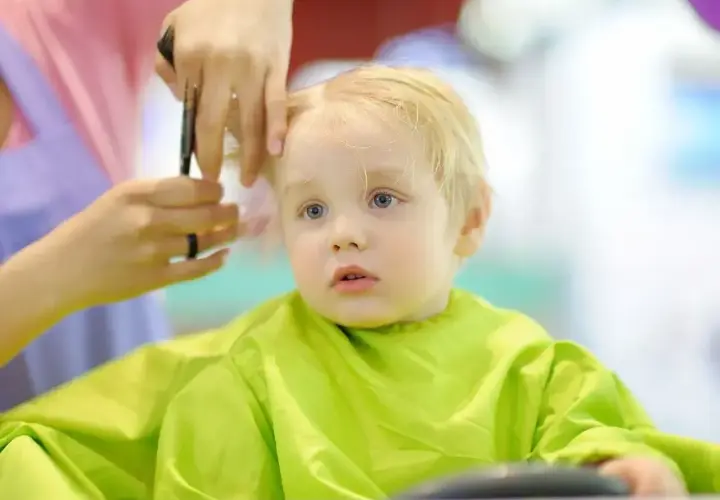 Mały chłopiec z blond włosami przeżywa swoje pierwsze strzyżenie dziecka. Fryzjerka delikatnie podcina mu włosy.