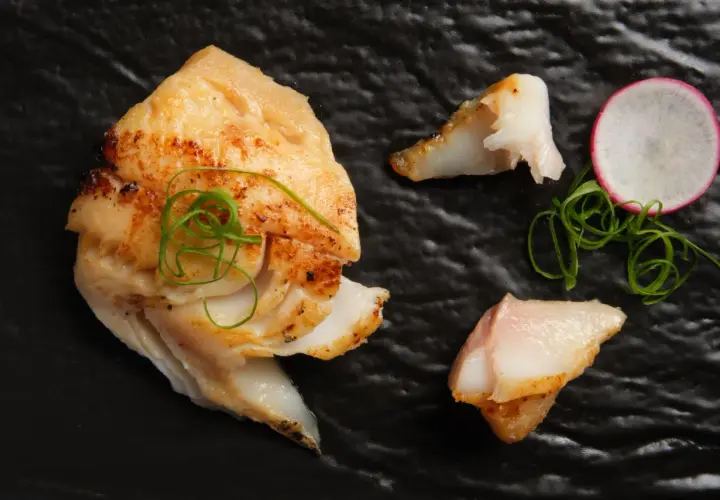 A beautifully grilled saikyo yaki fish fillet, garnished with green onions and a radish slice, rests on a black plate.