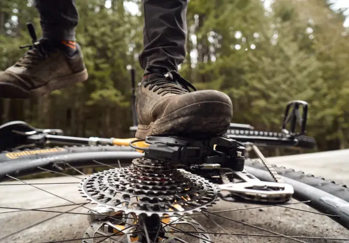 A mountain biker's muddy shoe rests on the SRAM T-Type derailleur and cassette of a bike lying on its side.