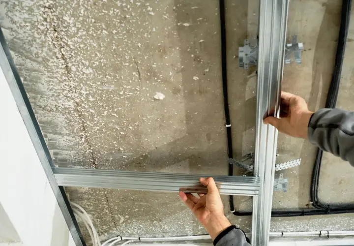 Ręce montują konstrukcję sufitu podwieszanego, łącząc profile aluminiowe. Widać kable i elementy mocujące.