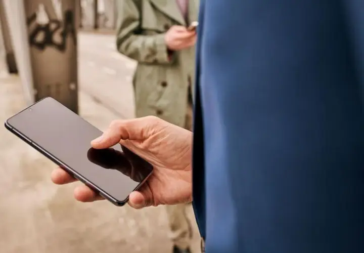 Dłoń trzyma smartfon z ciemnym ekranem. W tle druga osoba również korzysta z telefonu.
