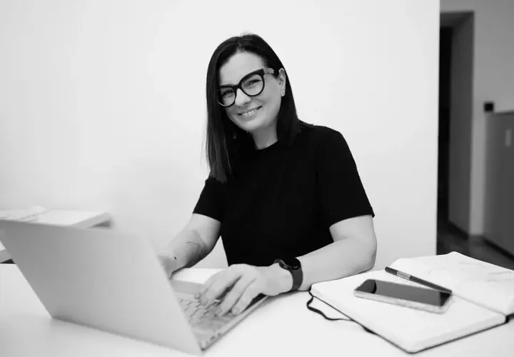 Kobieta w okularach pracuje na laptopie przy biurku z notatnikiem i telefonem.