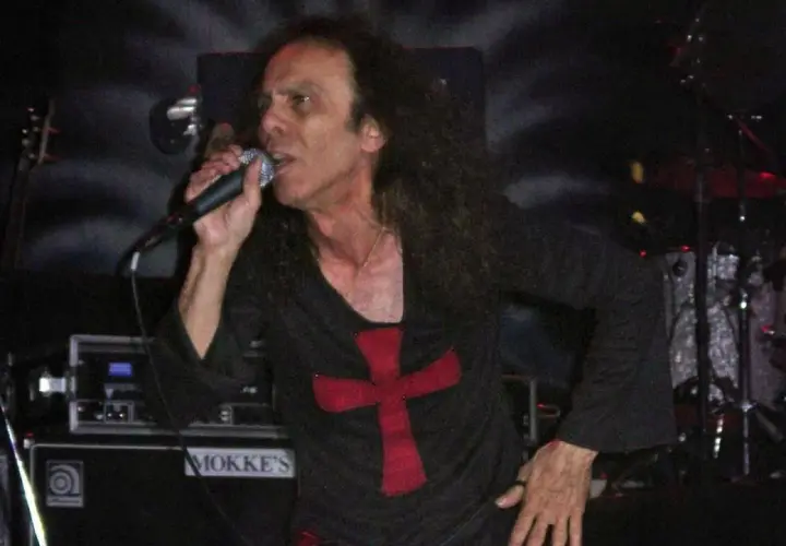 Ronnie James Dio zespół śpiewa do mikrofonu, ubrany w czarną koszulkę z czerwonym krzyżem.