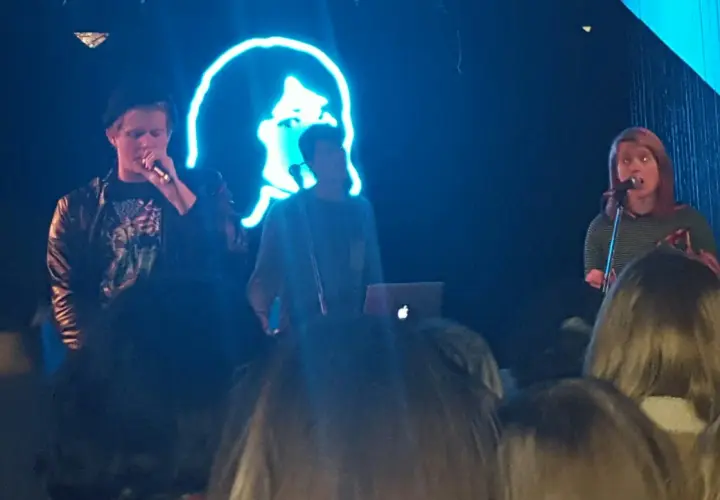 Koncert zespołu tv girl. Na scenie trójka artystów, w tle neonowa podobizna twarzy.