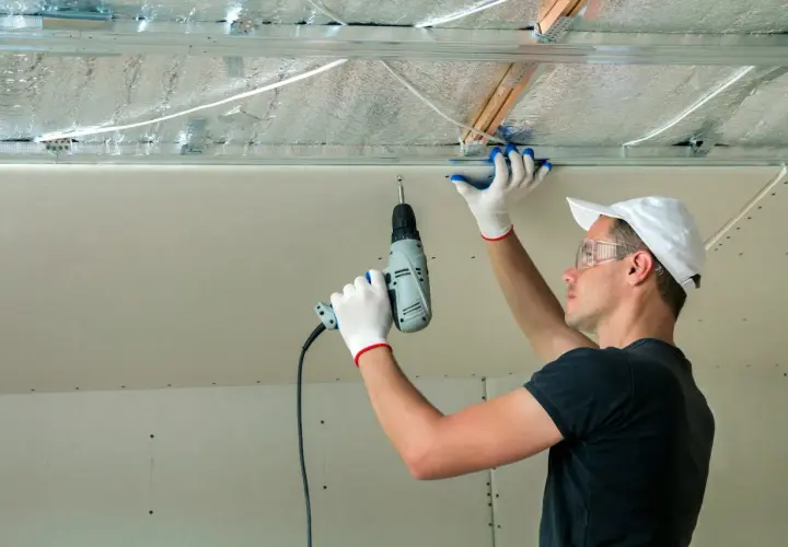 Un ouvrier installe des isolants minces sous un faux plafond en plaques de plâtre, utilisant une perceuse pour fixer les éléments.