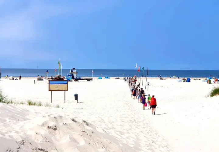 Menschen spazieren auf einem Holzsteg über den weiten, weißen nordsee strand. Der Himmel ist blau, das Meer ruhig.