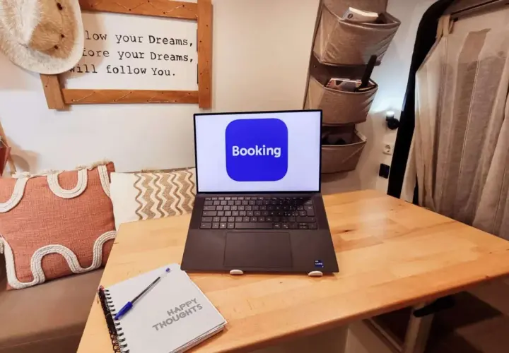 Laptop con logo Booking aperto su un tavolo di legno, accanto a un quaderno "Happy Thoughts". Pianifica il tuo prossimo viaggio con credito booking.