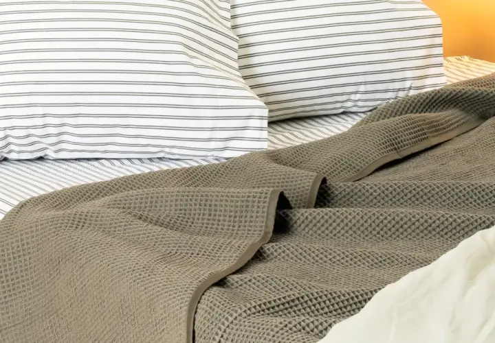 A textured, grey blanket is draped at the end of a bed, over striped pillows and sheets.