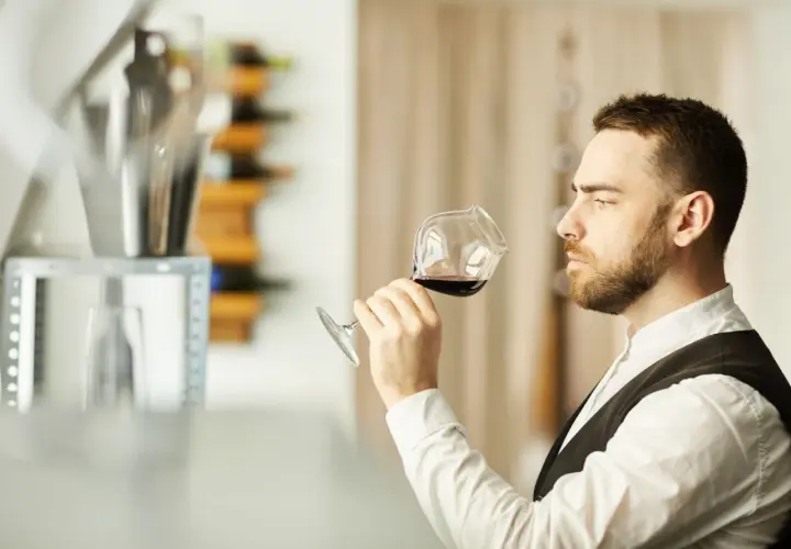 Mężczyzna w białej koszuli i kamizelce, sommelier, kto to? Wącha kieliszek czerwonego wina, oceniając jego aromat.