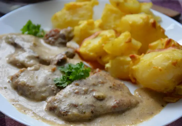 Pyszny karczek w sosie chrzanowym z pieczonymi ziemniakami. Danie ozdobione natką pietruszki.