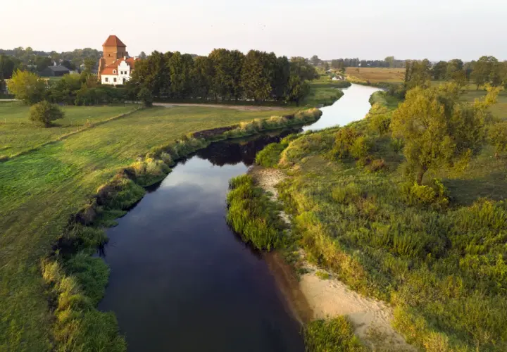 Malownicza wieś nad Liwcem, z charakterystycznym dworem i wieżą, otoczona zielonymi łąkami i drzewami. Rzeka Liwiec meandruje przez krajobraz.