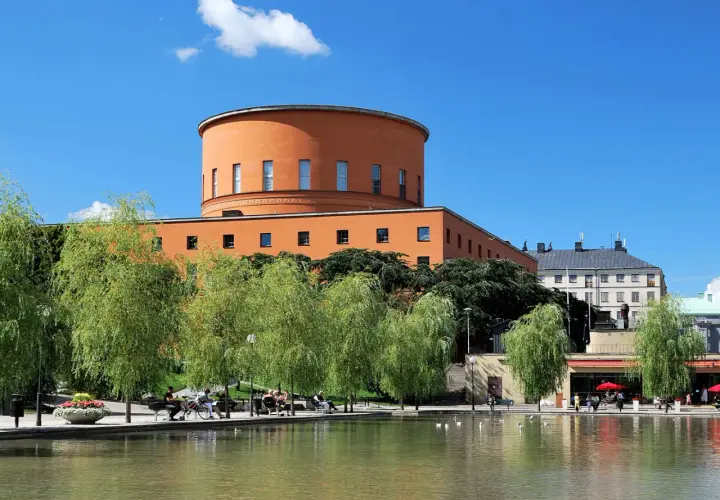 En vacker stadsvandring i Stockholm på egen hand. En orange, rund byggnad reser sig bakom grönskande träd vid en damm.
