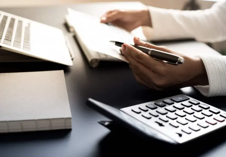 Ręce trzymają długopis nad dokumentami, obok kalkulator i laptop. Analiza danych, by dowiedzieć się, ile zarabia dietetyk.