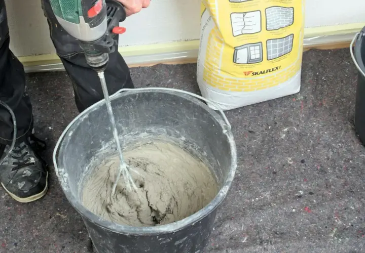 Personen använder en borrmaskin för att blanda betong i en hink. En gul säck med Skalflex-märkning står bredvid.