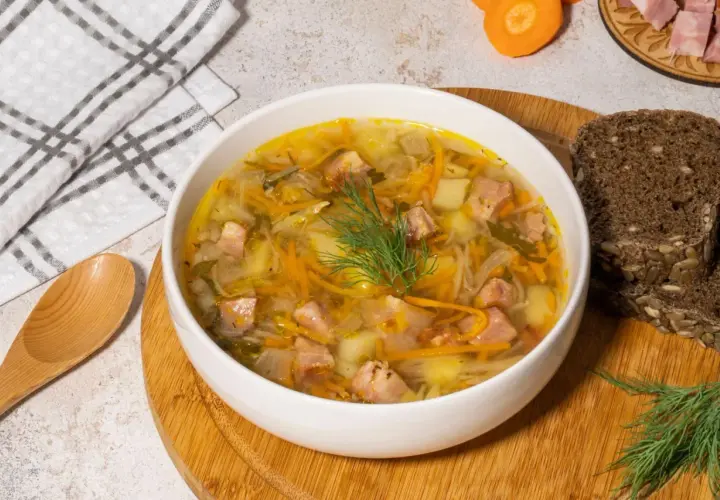 Aromaticzny kapuśniak ze słodkiej kapusty z kawałkami mięsa, ziemniakami i marchewką, udekorowany koperkiem. Obok kromki ciemnego chleba.