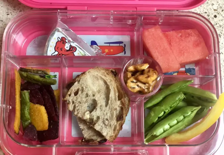 A pink bento box with watermelon, peas, pretzels, bread, and veggie sticks. Add an ice pack to keep this bento box cold.