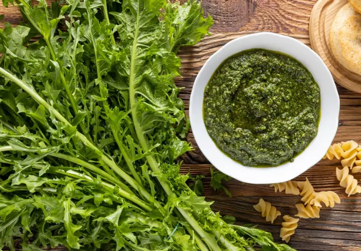 Un mazzo di **tarassaco foglie** fresche accanto a una ciotola di pesto verde brillante e pasta fusilli su un tavolo di legno rustico.