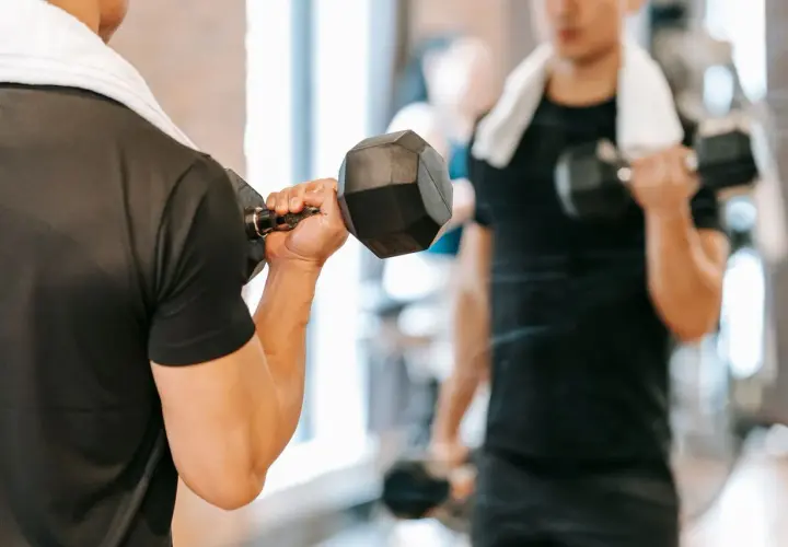 Mężczyzna ćwiczy biceps z hantlem, zaczynając dzień od treningu na czczo. W tle widać jego odbicie w lustrze.