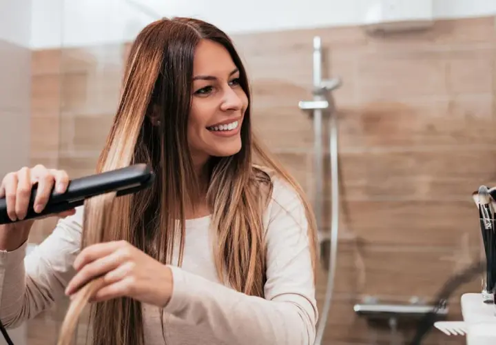 Mujer sonriendo mientras se alisa el pelo con una plancha, buscando el secreto de como alisar el pelo para siempre.